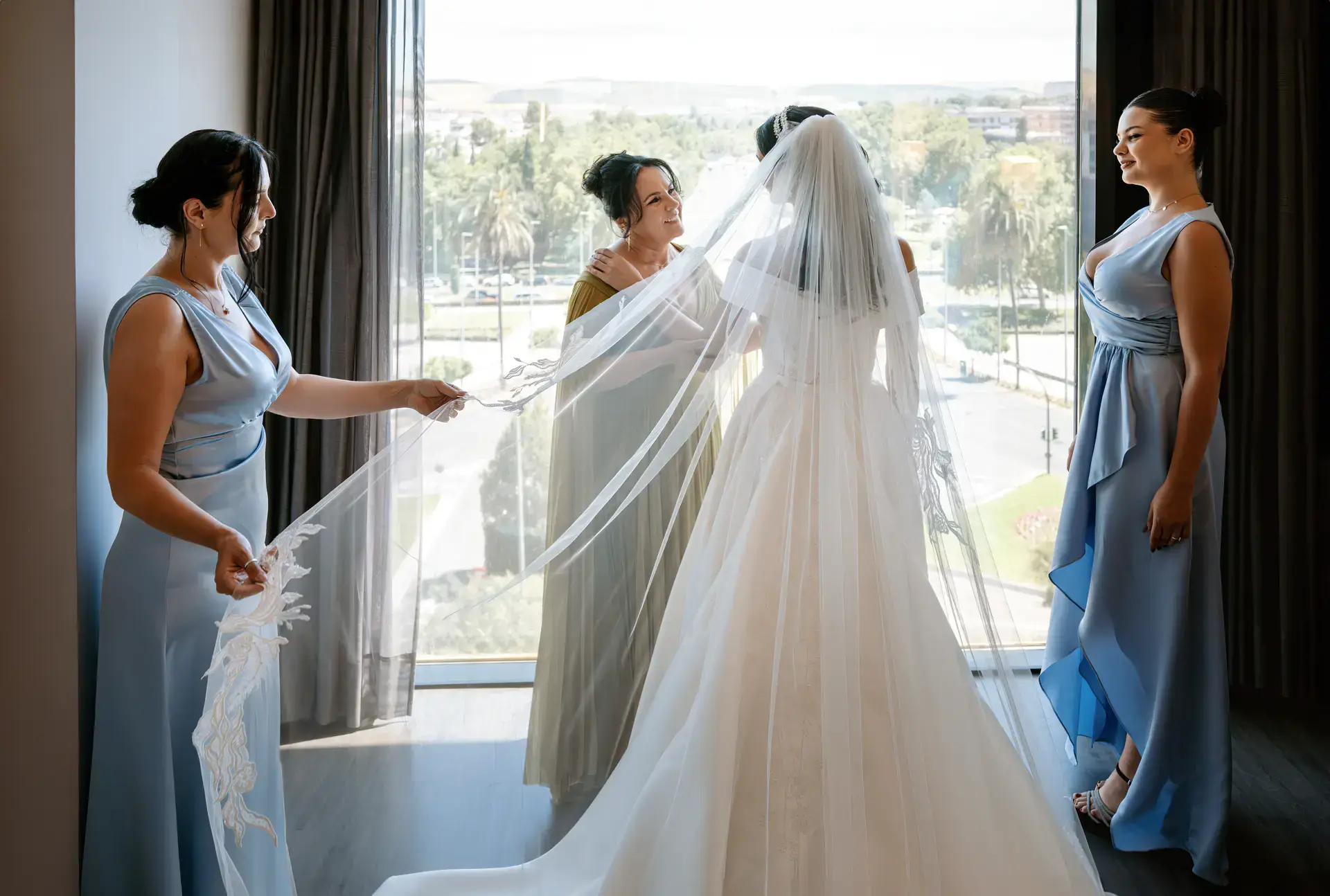 Las damas de honor ayudan con el velo a la novia junto al ventanal