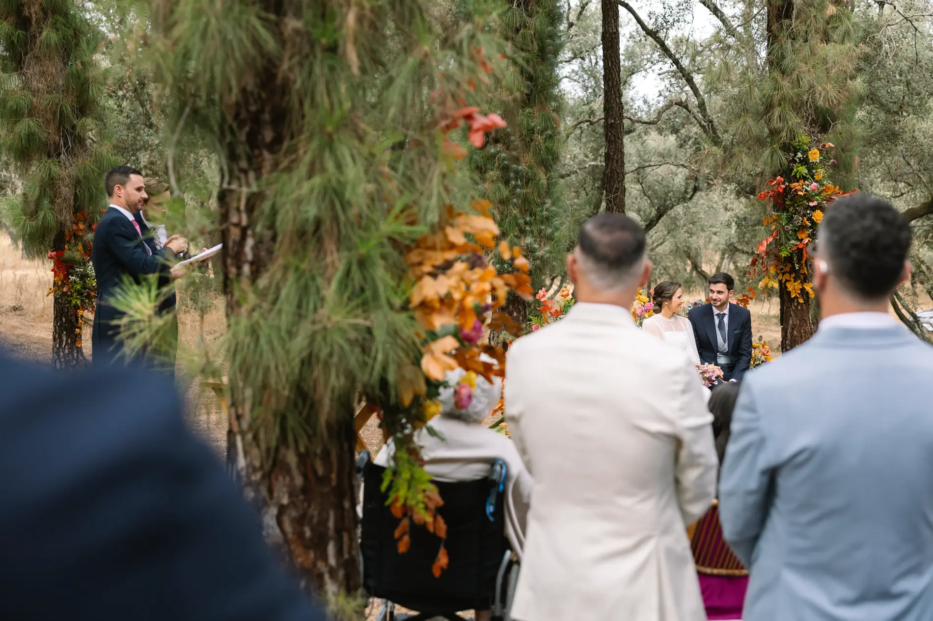 Vista de la ceremonia entre los árboles desde detrás de los invitados, en Casa Colorada, Badajoz