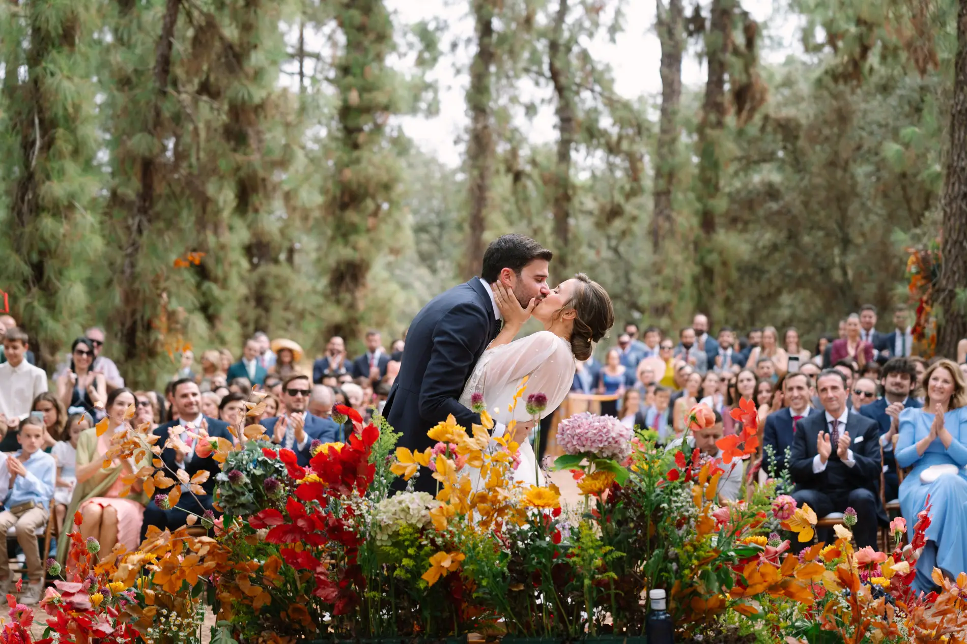 Álvaro y María se besan tras un gran arreglo floral de tonos naranjas y rosas durante la ceremonia al aire libre en el Marquesado de Casa Colorada, con los invitados al fondo.