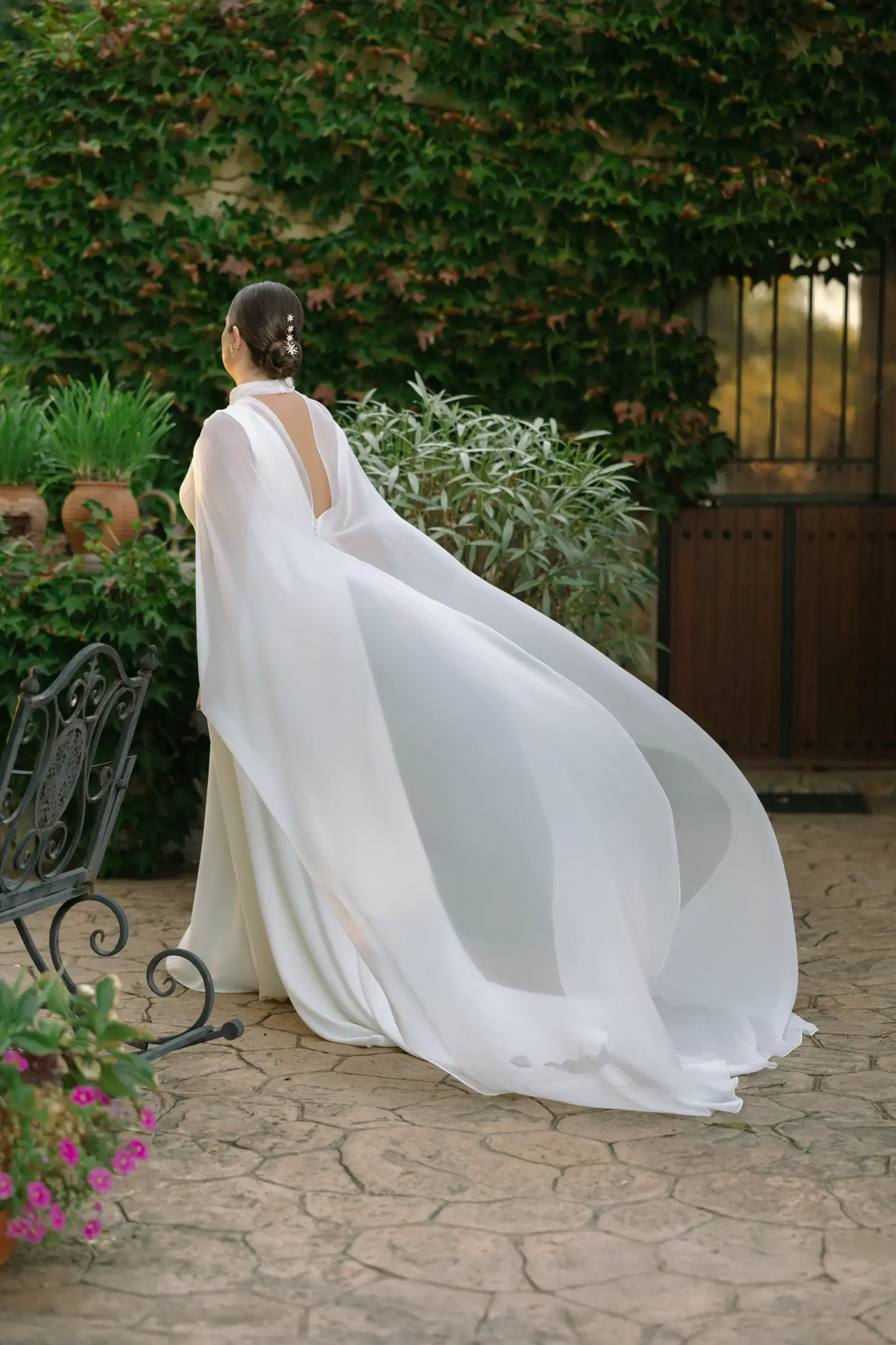La novia con capa al viento en un patio con flores