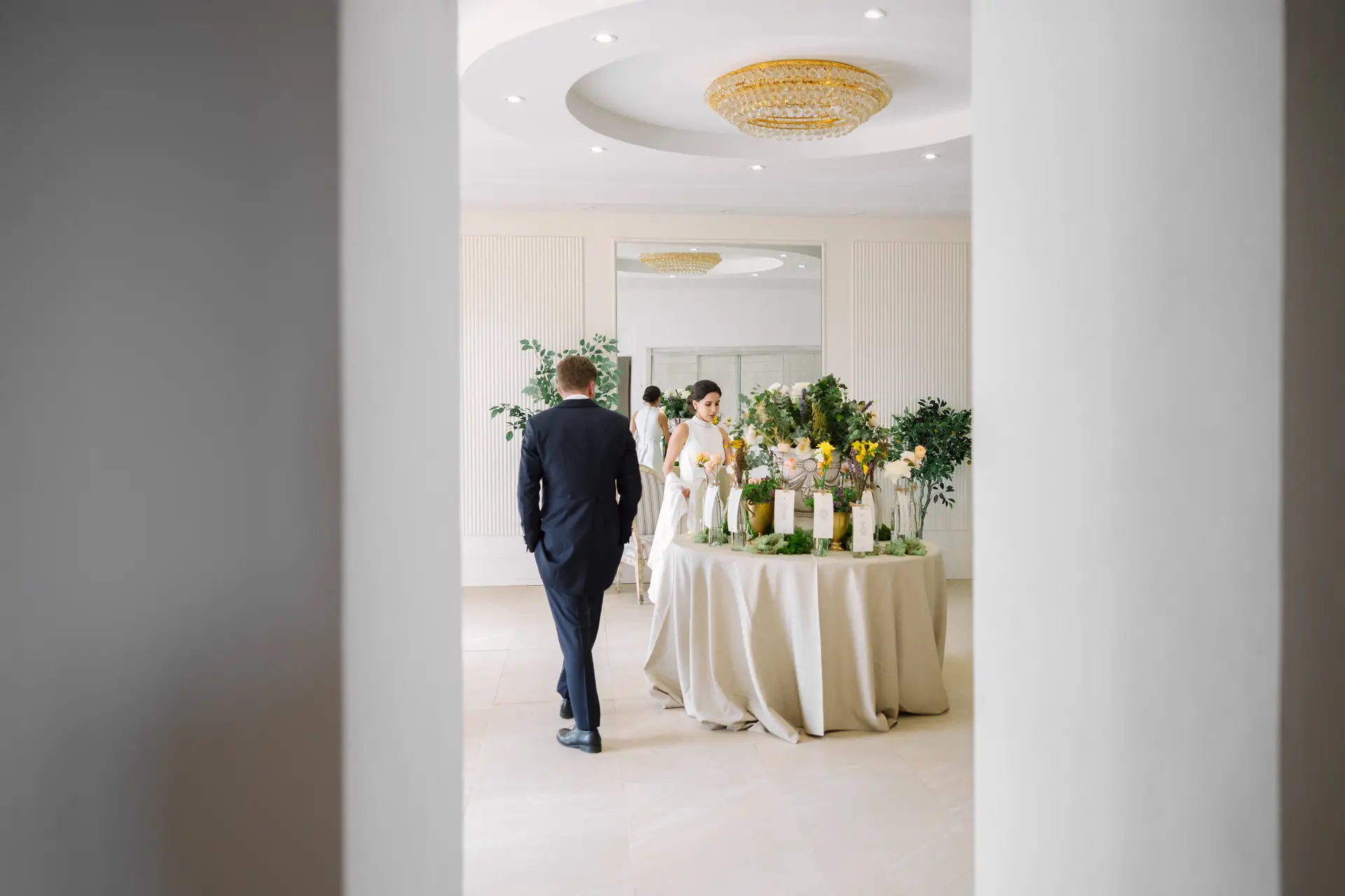 El novio camina hacia una mesa de flores en un salón blanco