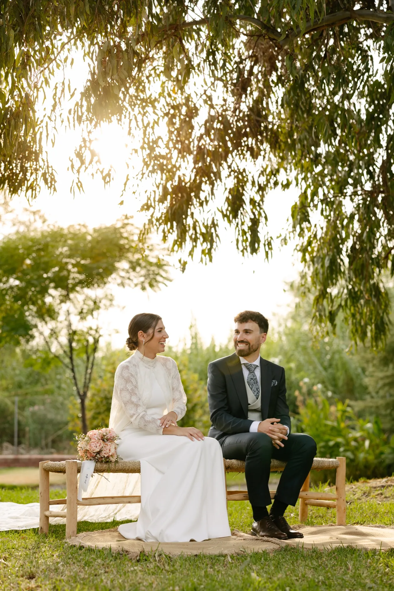 Los novios sonríen sentados en un banco de madera durante la ceremonia, bajo un árbol al atardecer