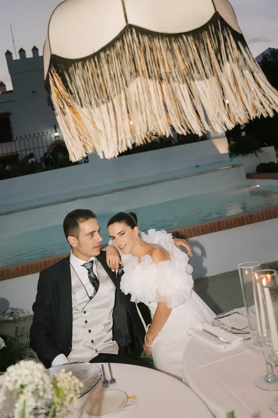 Los novios sentados junto a una piscina bajo una lámpara de flecos