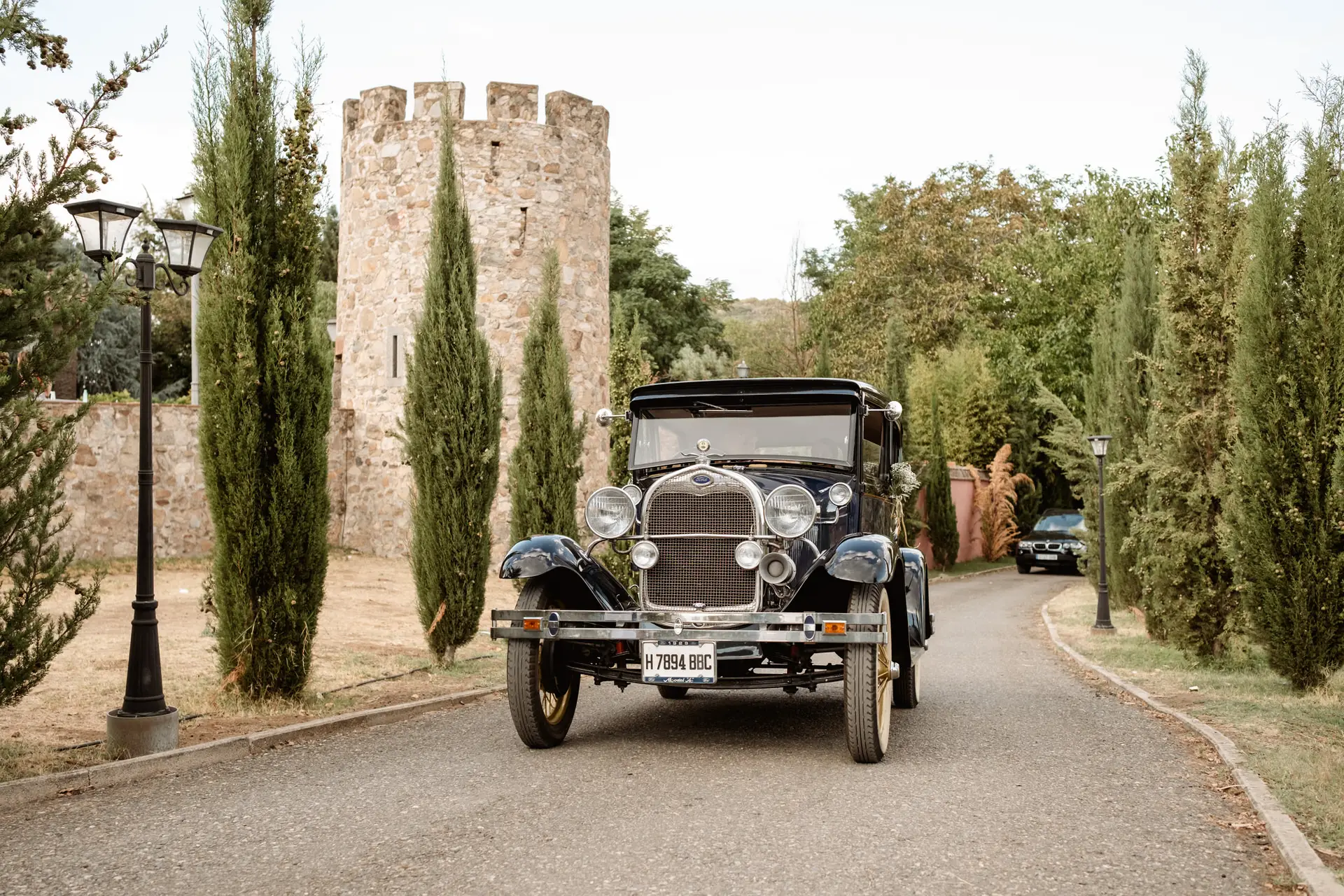 El coche clásico de la novia llega entre cipreses, con la torre del castillo al fondo