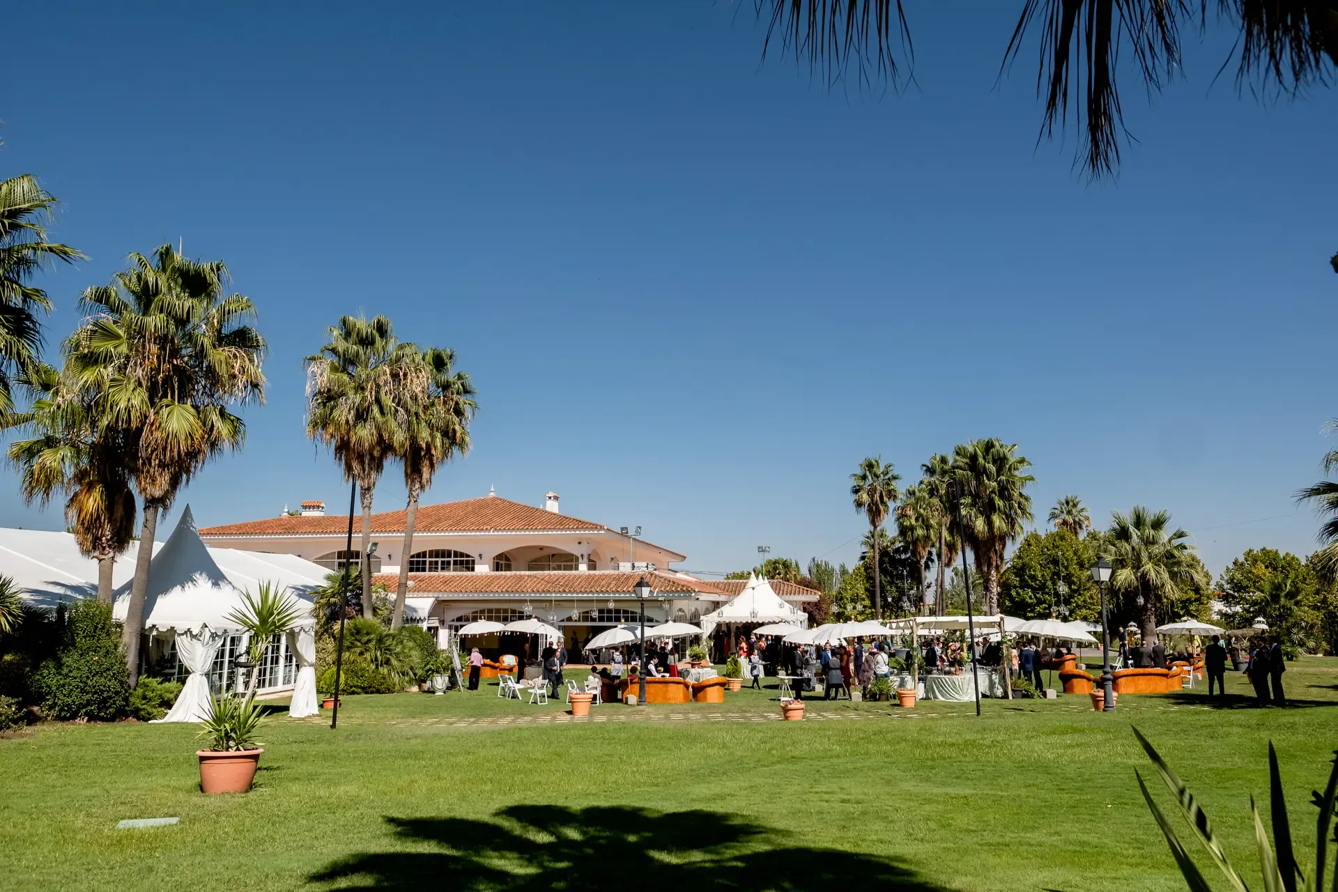 Cóctel bajo carpas blancas y palmeras en los jardines del Golf Guadiana (Badajoz), con el edificio del club al fondo