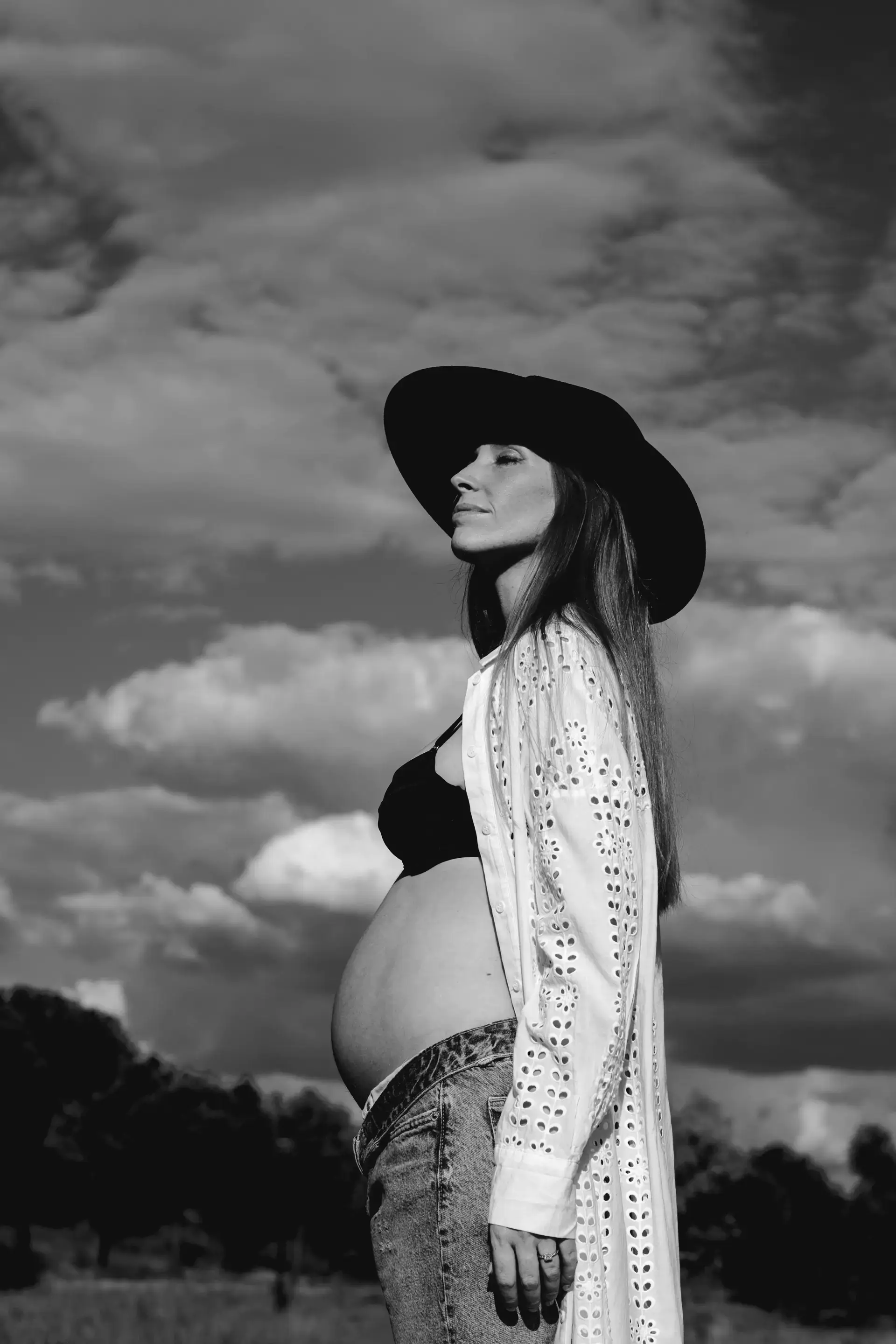 Mujer embarazada de perfil con sombrero ante un cielo de nubes, en blanco y negro