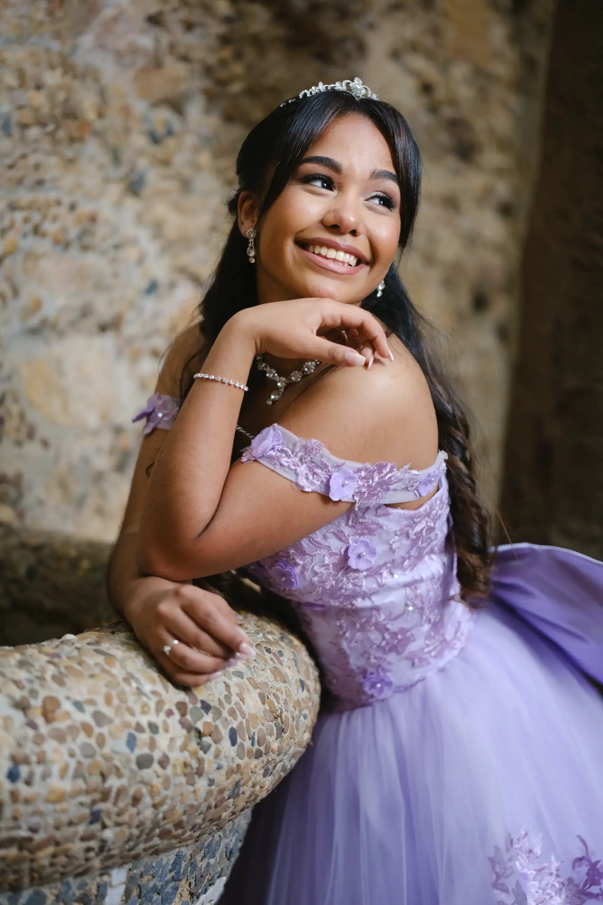 Quinceañera con vestido lila de princesa y tiara junto a un muro de piedra