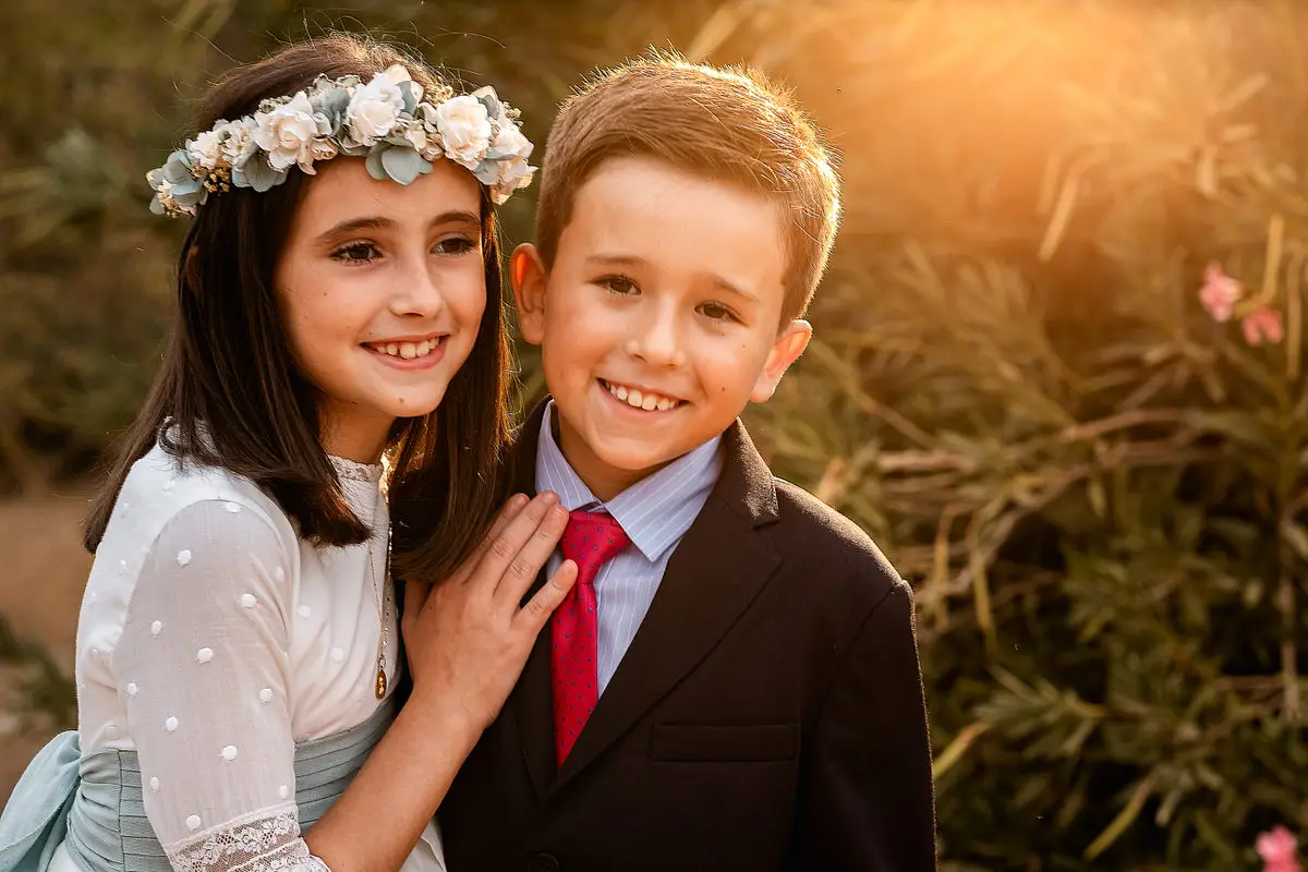 Niña con corona de flores y niño con traje de comunión abrazados al atardecer