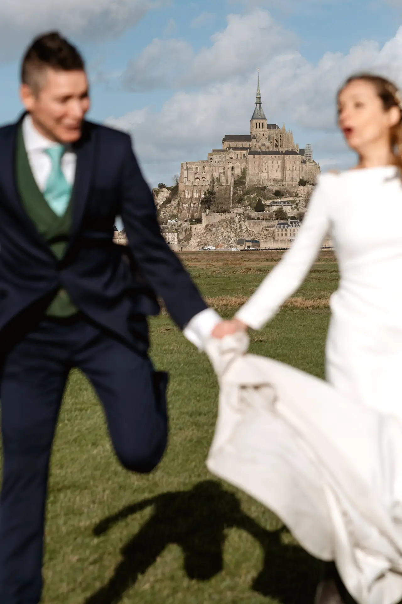Los novios corren de la mano con el Mont Saint-Michel al fondo
