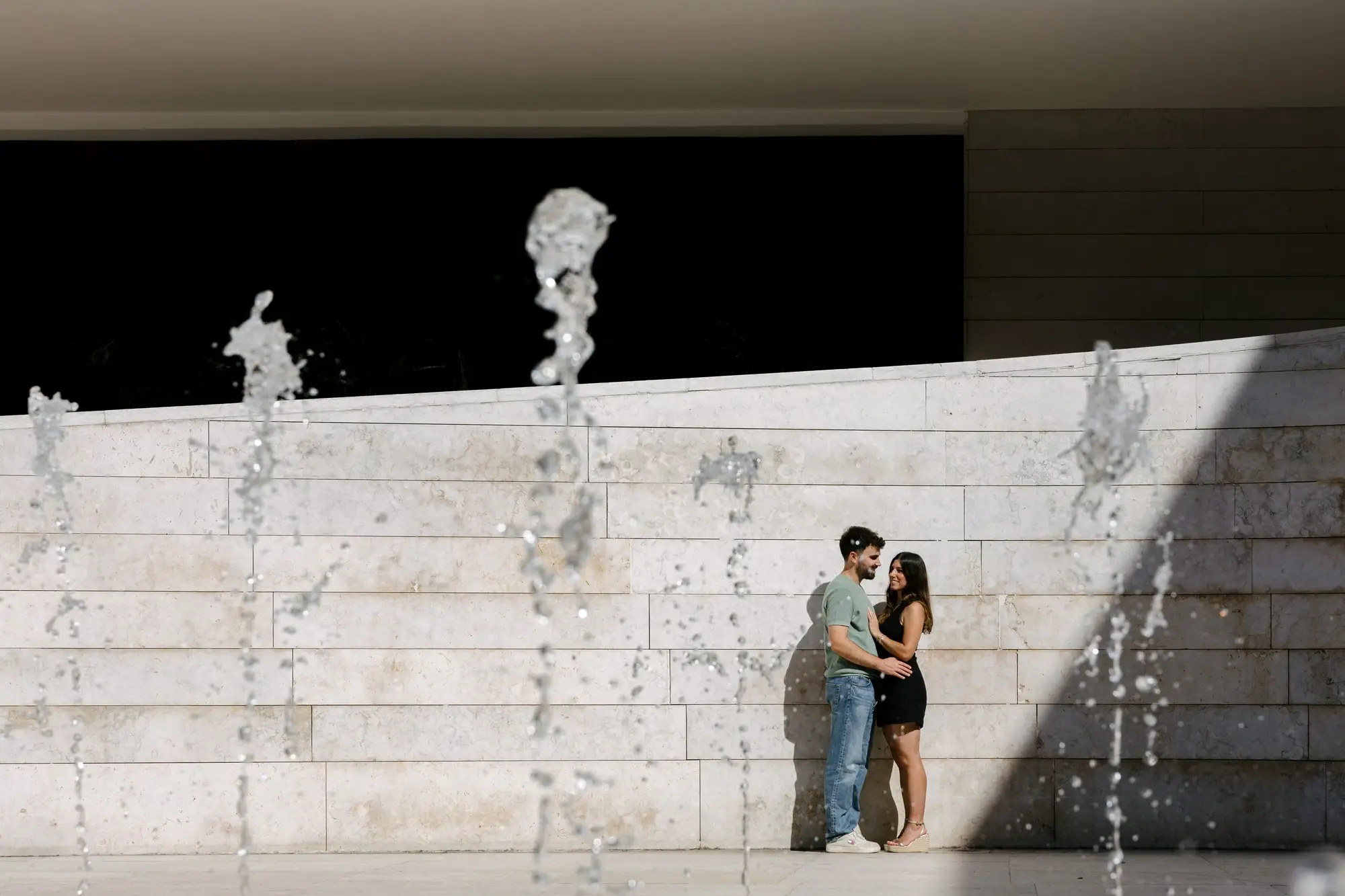 Marina y Juan Antonio abrazados junto a un muro de piedra con chorros de agua, en Lisboa