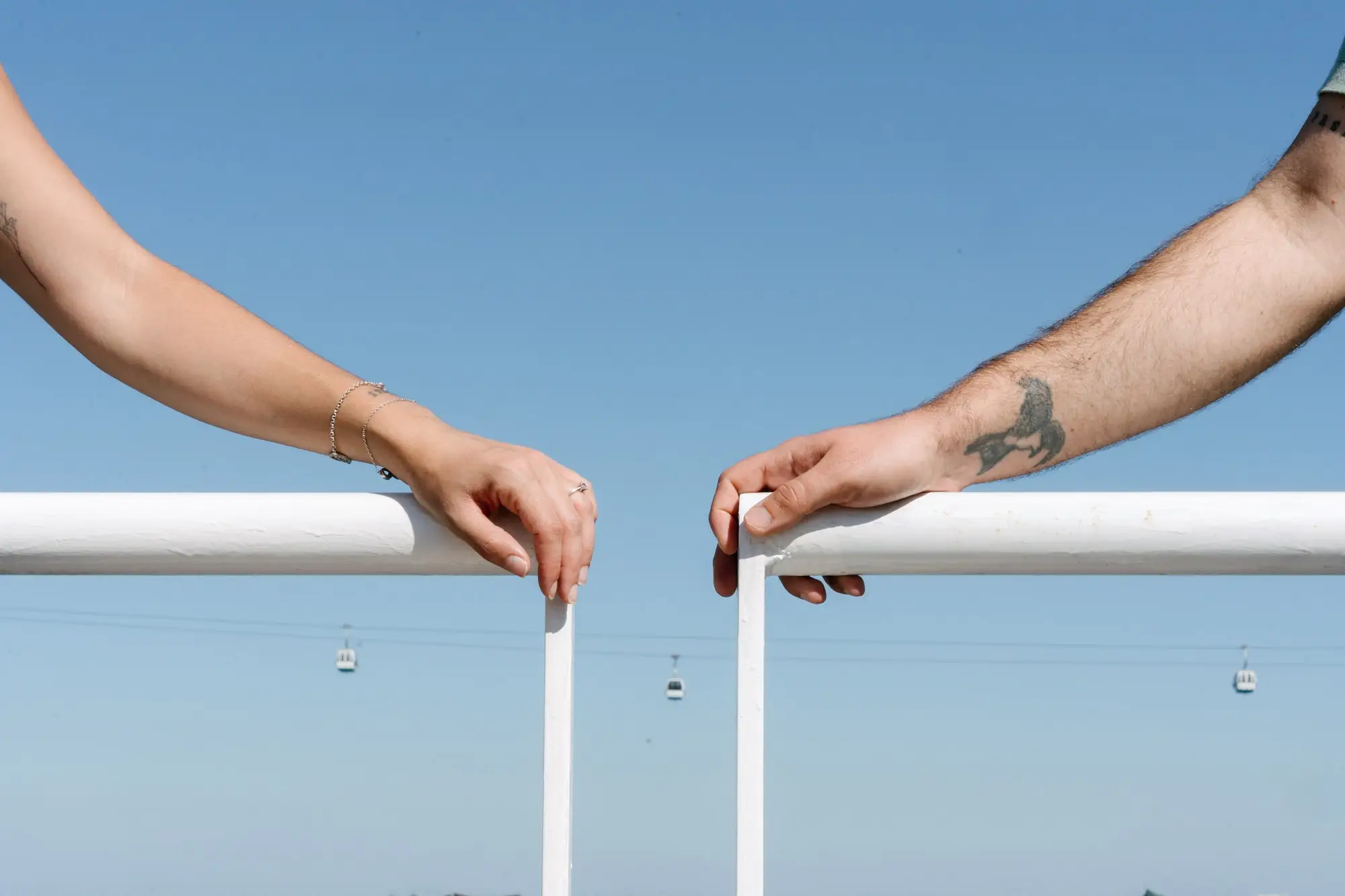Las manos de Marina y Juan Antonio a punto de tocarse sobre una barandilla blanca, con el teleférico al fondo, en Lisboa