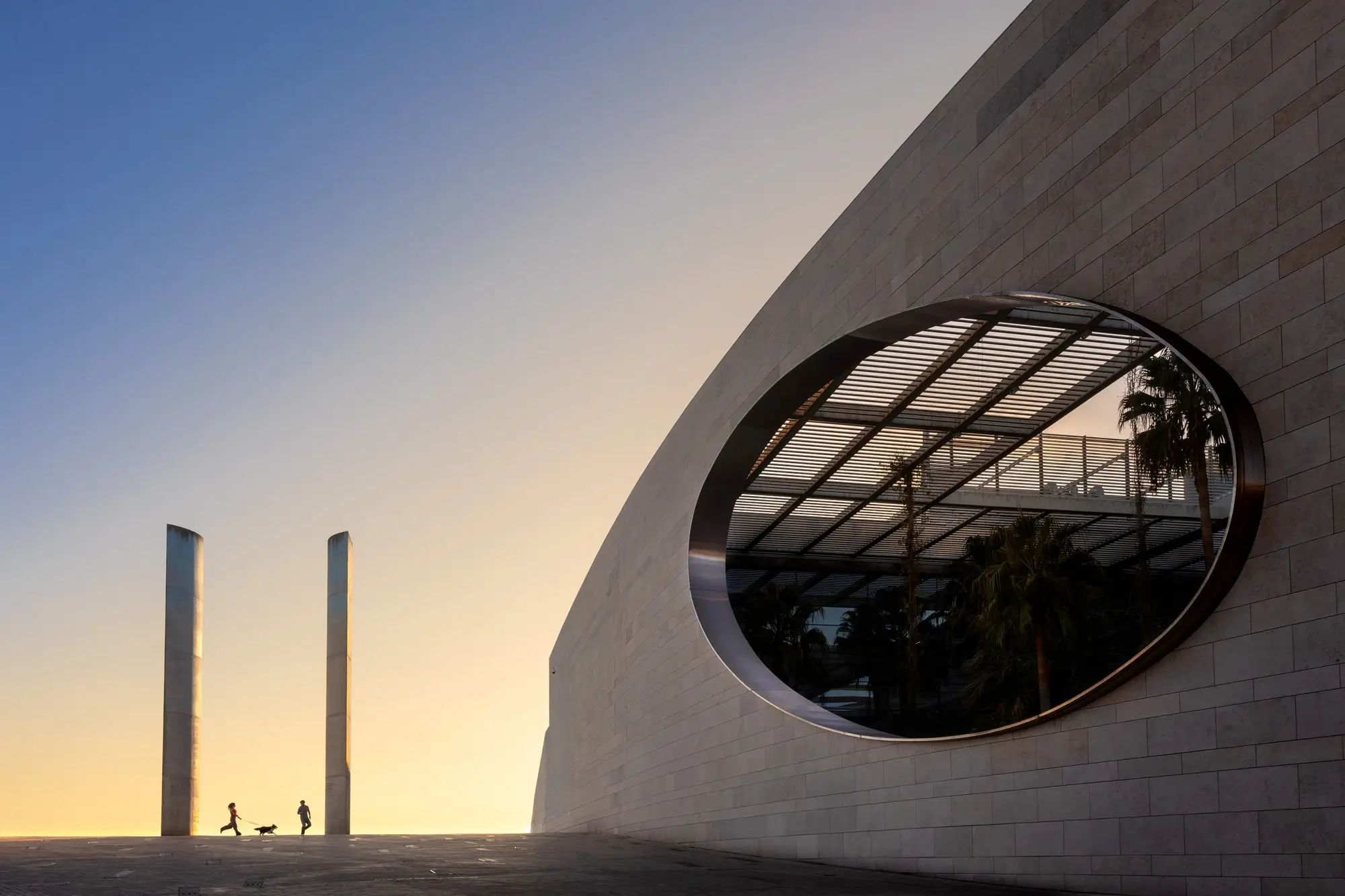 Muro curvo con una ventana ovalada y dos columnas altas al atardecer, con la pareja diminuta al fondo, en Lisboa