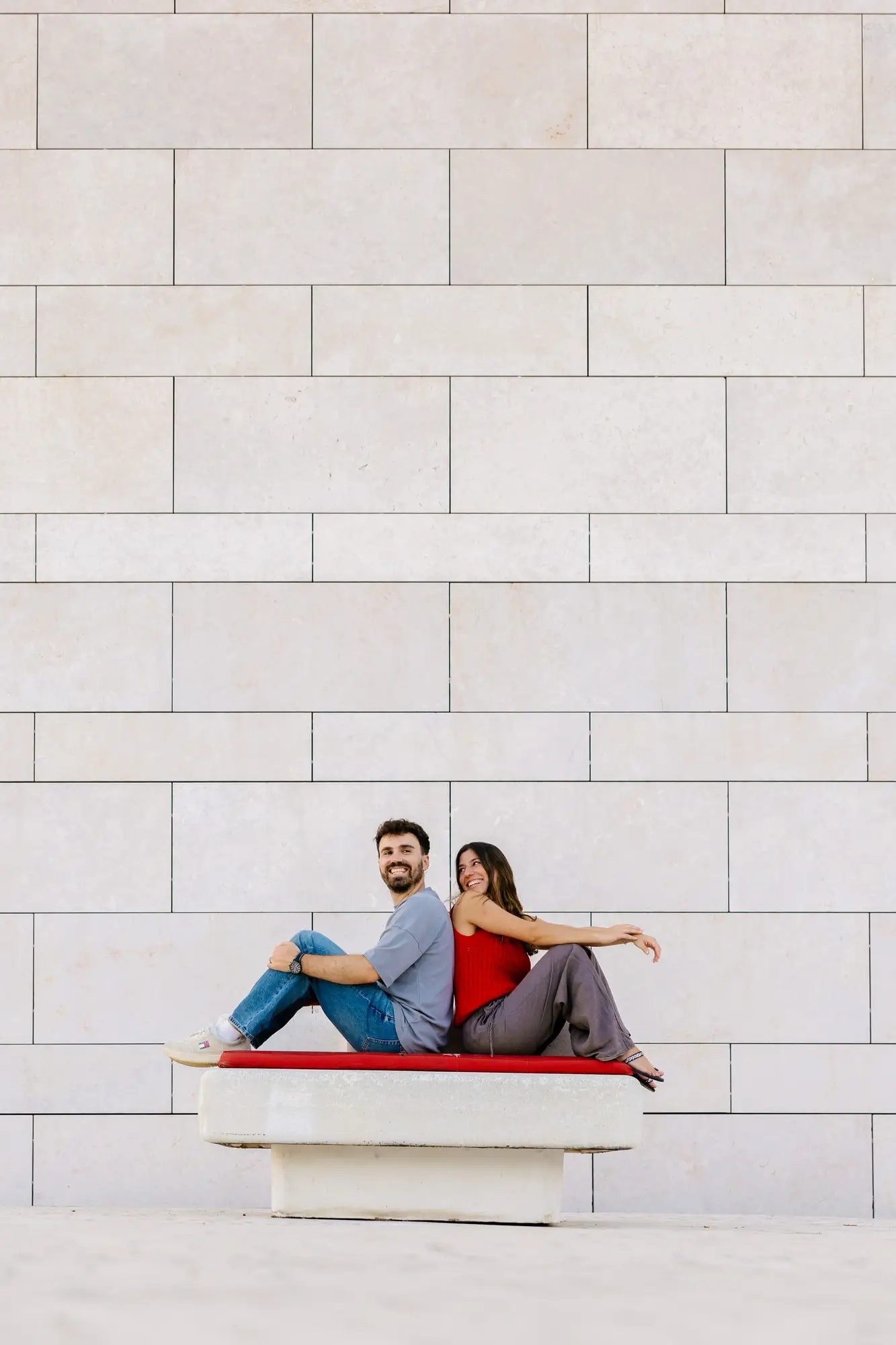 Marina y Juan Antonio sentados en un banco rojo frente a un muro de piedra clara, en Lisboa