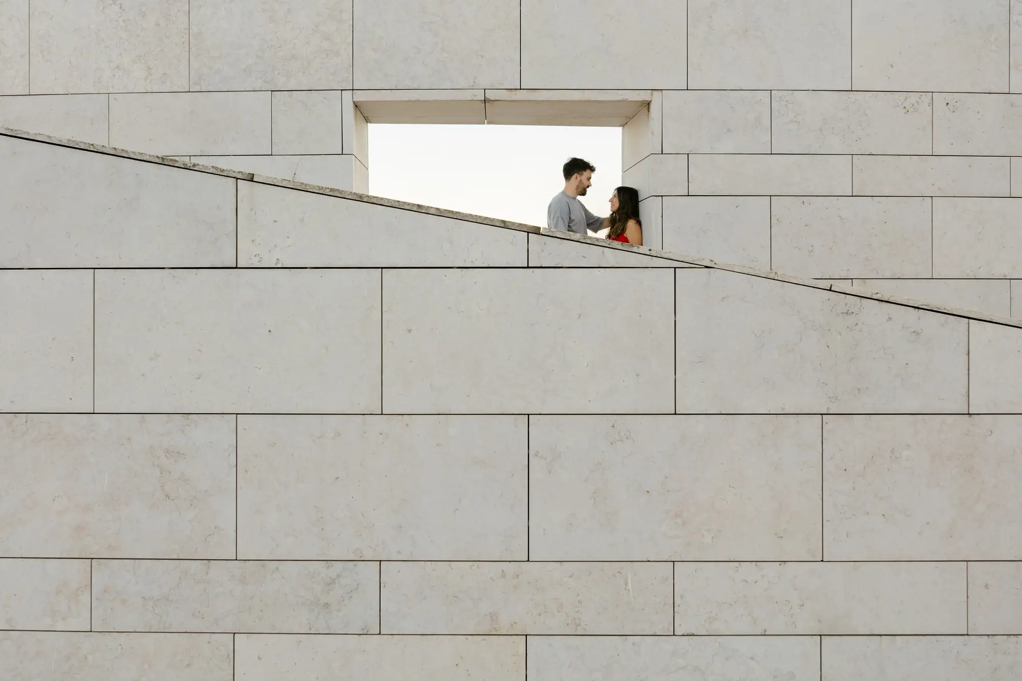 Marina y Juan Antonio asomados a una ventana pequeña en un muro de piedra cruzado por una diagonal, en Lisboa