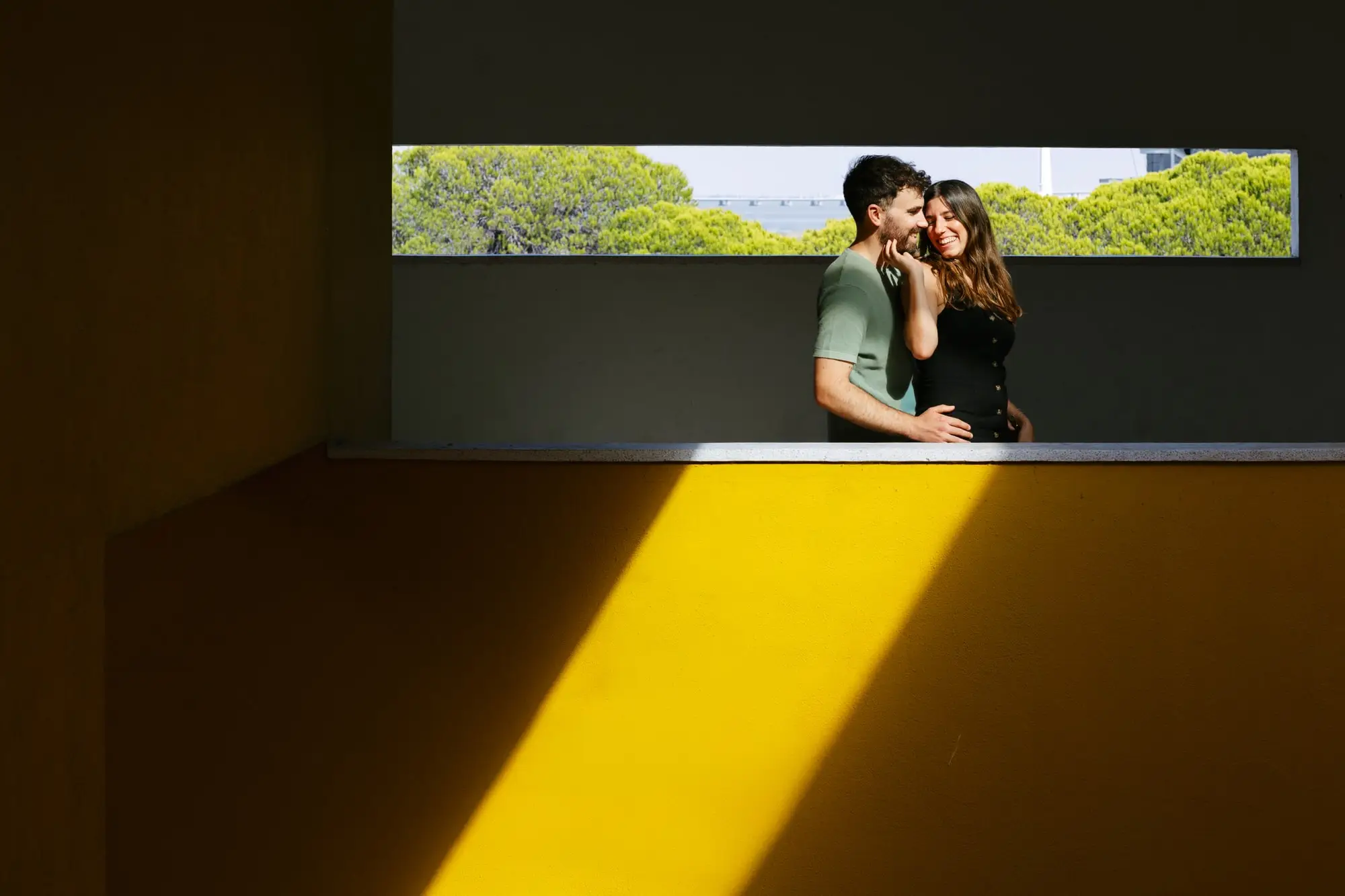 Marina y Juan Antonio abrazados en una ventana alargada, sobre una pared amarilla cortada por una franja de sol, en Lisboa