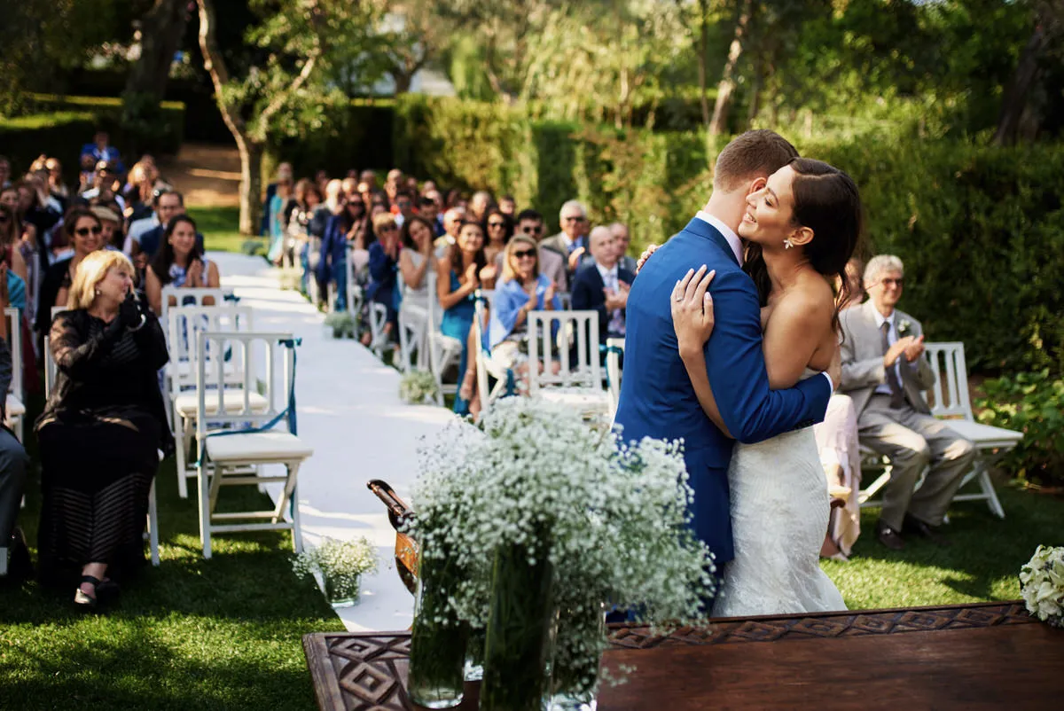 Novios abrazándose emocionados tras su ceremonia de boda en un jardín al aire libre con invitados aplaudiendo en el fondo