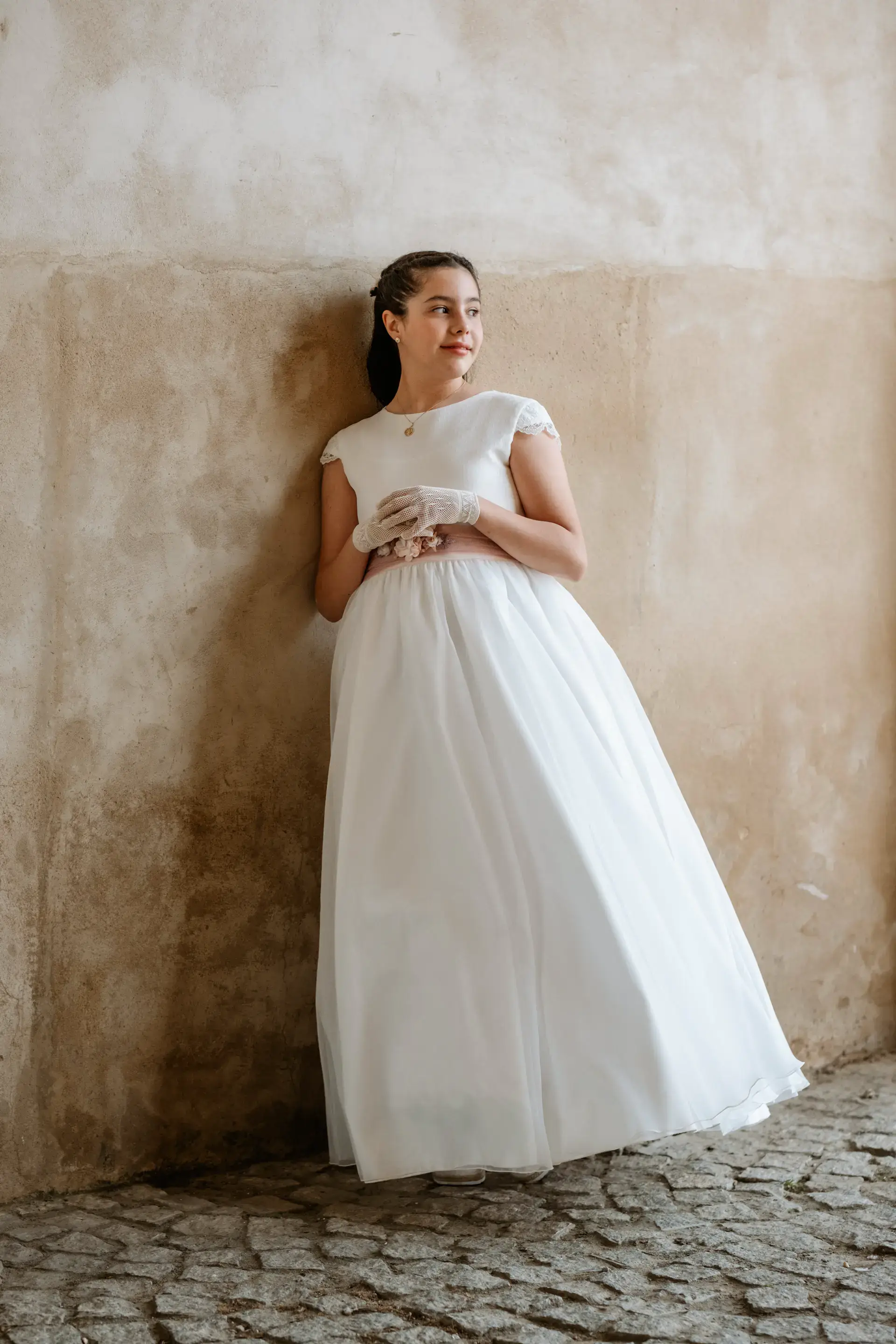 Niña con vestido de comunión blanco apoyada en un muro