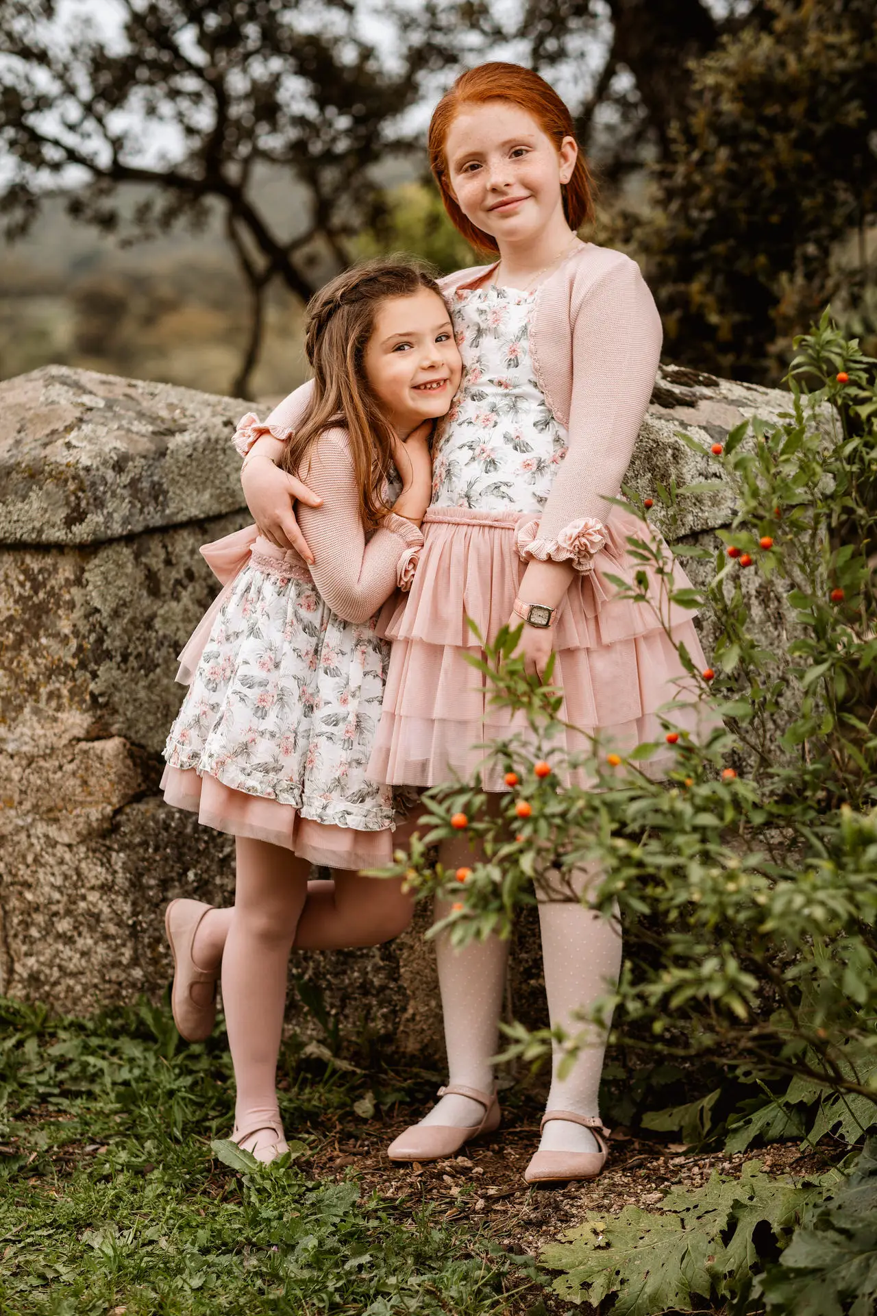 Dos hermanas con vestidos de flores abrazadas junto a un muro de piedra